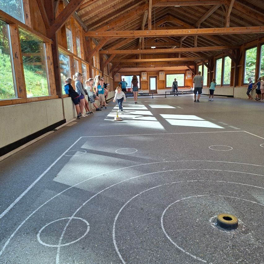 Eine Gruppe von Kindern und Erwachsenen spielt eine Partie Kegelspiel in einem holzgedeckten Innenbereich. Das Spiel ist auf einem Betonboden mit Kreidemarkierungen ausgelegt.