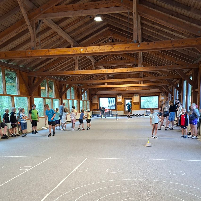 Eine Gruppe von Kindern und Erwachsenen versammelt sich in einem großen, von Holzbalken durchzogenen Innenbereich, möglicherweise einer Sporthalle. Sie stehen um ein kreisförmiges Muster auf dem Boden herum, wobei einige Personen in Aktivitäten verwickelt sind. Der Raum hat große Fenster und eine hohe Decke.
