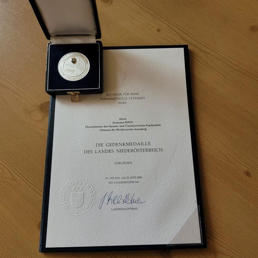 Ein Zertifikat und eine Medaille für lebenslange Dienste, präsentiert in einer Box auf einer Holzoberfläche. Das Zertifikat ist von der Regierung von Niederösterreich.