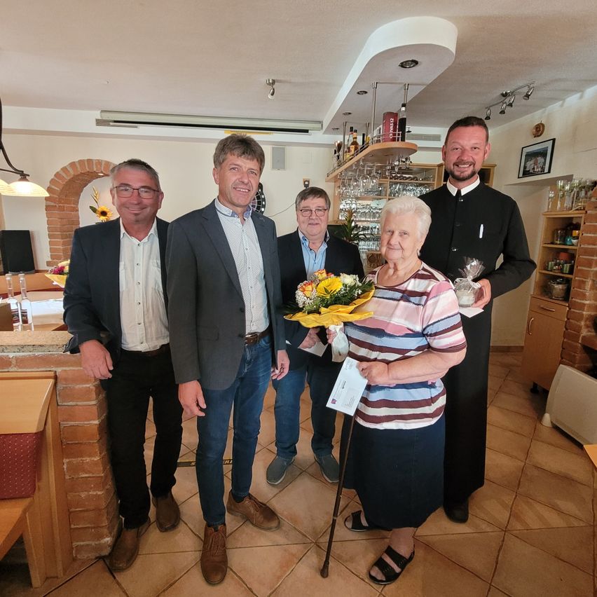 Five individuals stand together in a room, smiling for a photo. A woman holds a bouquet of flowers and a paper, while a man in a black robe stands behind her.
