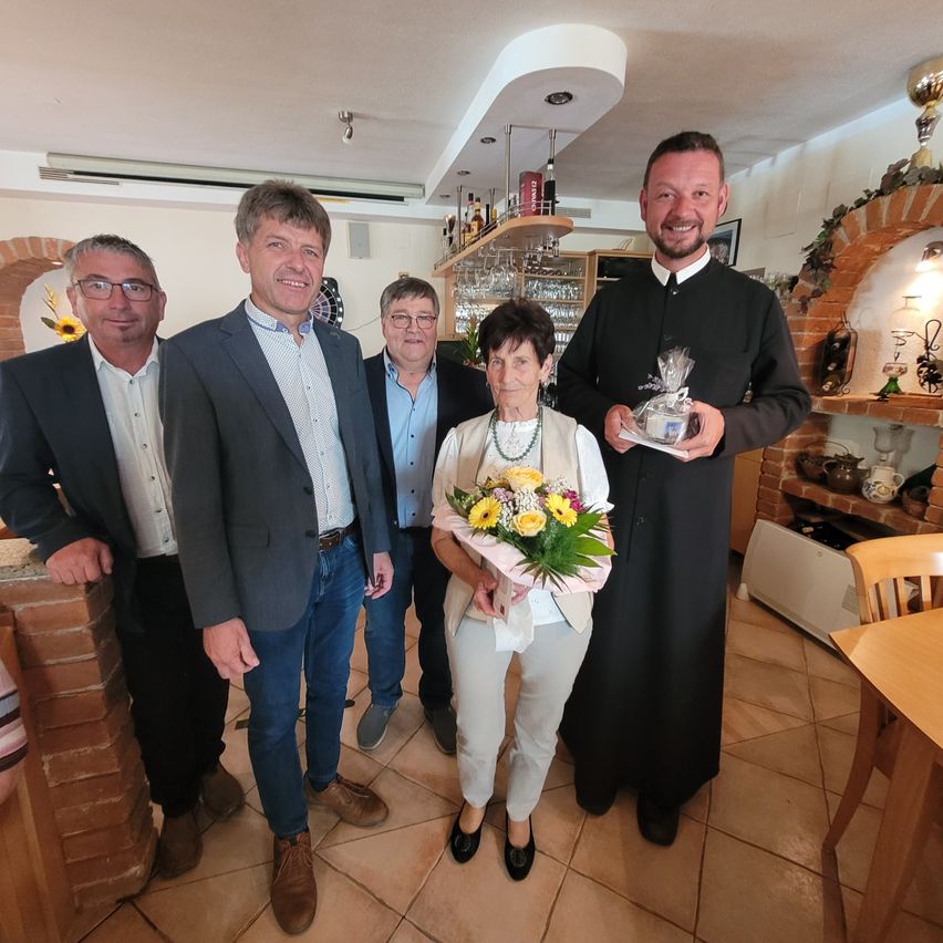 A group of people stands in a room with a priest holding a gift. A woman in the center holds a bouquet of flowers. Behind them is a shelf with bottles and glasses.
