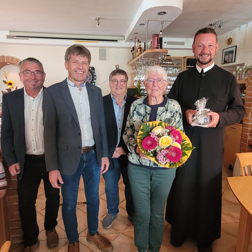 Five people stand together in a room, smiling for a photo. A woman in the center holds a bouquet of flowers. Behind her, a man in a black robe holds a wrapped gift.