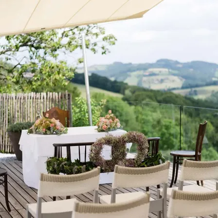 Eine Hochzeitszeremonie auf einer Holzterrasse mit Blick auf eine malerische Berglandschaft. Der Altar ist mit Blumenarrangements geschmückt, und Stühle sind davor aufgestellt.