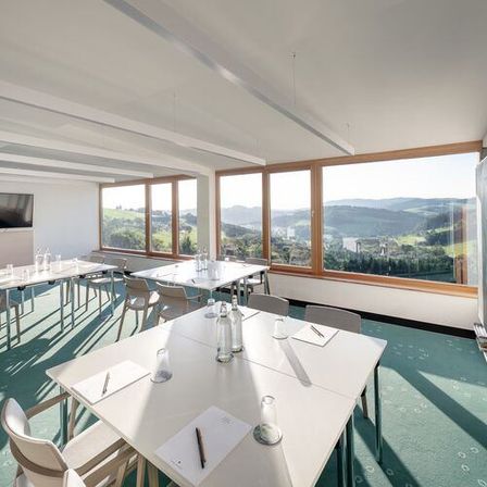 Ein leerer Konferenzraum mit weißen Wänden, grünem Teppich und großen Fenstern mit Blick auf eine Berglandschaft. Es gibt mehrere Tische und Stühle im Raum, eine Tafel und ein Monitor an der Wand.