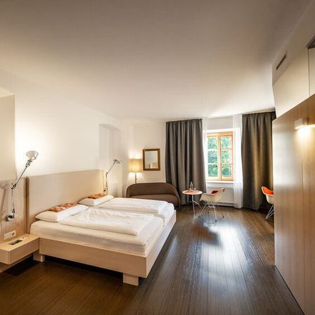 Ein Hotelzimmer mit zwei Betten, einer Couch, einem Tisch und einem Fenster mit Vorhängen. Das Zimmer hat Holzböden und weiße Wände.