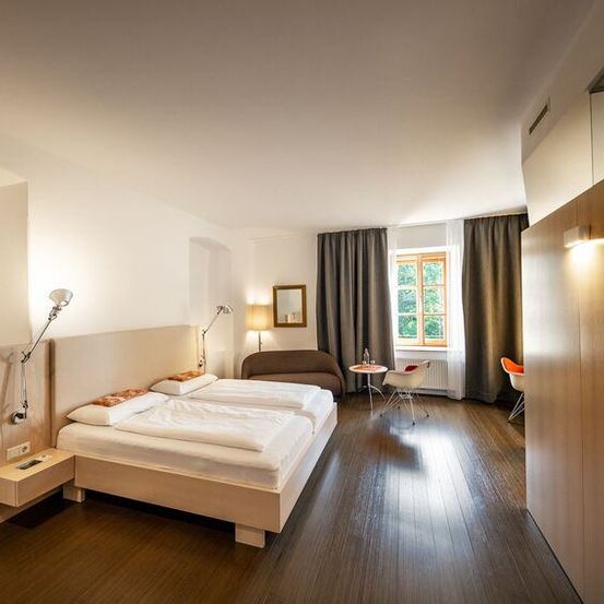 Ein Hotelzimmer mit zwei Betten, einer Couch, einem Tisch und einem Fenster mit Vorhängen. Das Zimmer hat Holzböden und weiße Wände.