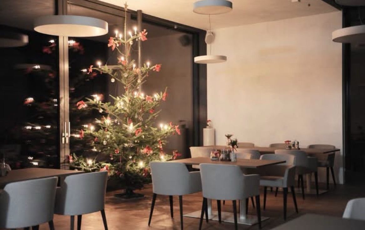 Ein warm beleuchteter Weihnachtsbaum steht in einem gemütlichen Restaurant, umgeben von Tischen und Stühlen, und schafft eine festliche Atmosphäre.