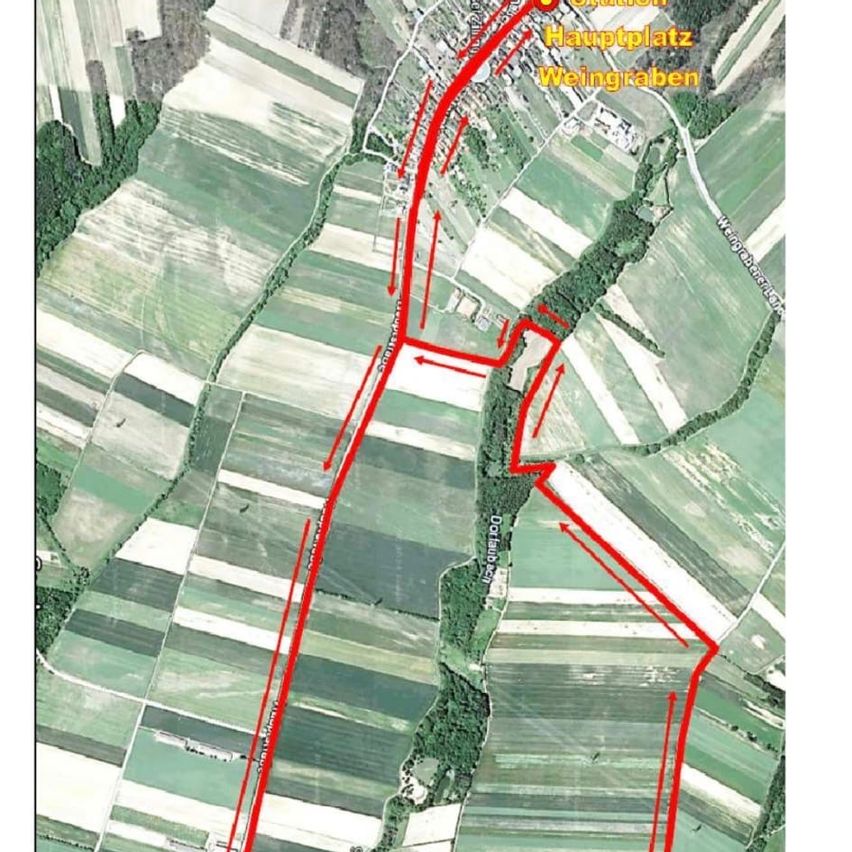 Luftaufnahme einer ländlichen Landschaft mit roten Linien, die Routen markieren. Grüne Felder sind durch Straßen getrennt. Oben befindet sich eine zentrale Beschriftung 'Hauptplatz Weingraben' und eine weitere Beschriftung 'Weingraben'.