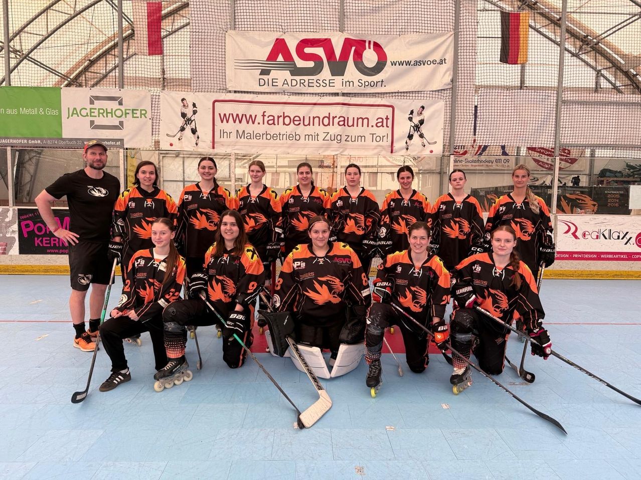 Eine Gruppe weiblicher Hockeyspielerinnen in orangen und schwarzen Trikots posiert für ein Foto auf der Indoor-Eisbahn.
