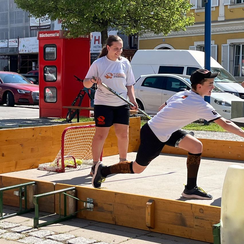 Zwei Kinder spielen ein Spiel Straßenhockey, wobei ein Kind läuft und das andere in einem Außenbereich mit einer Holzumzäunung und einer roten Telefonzelle zuschaut.