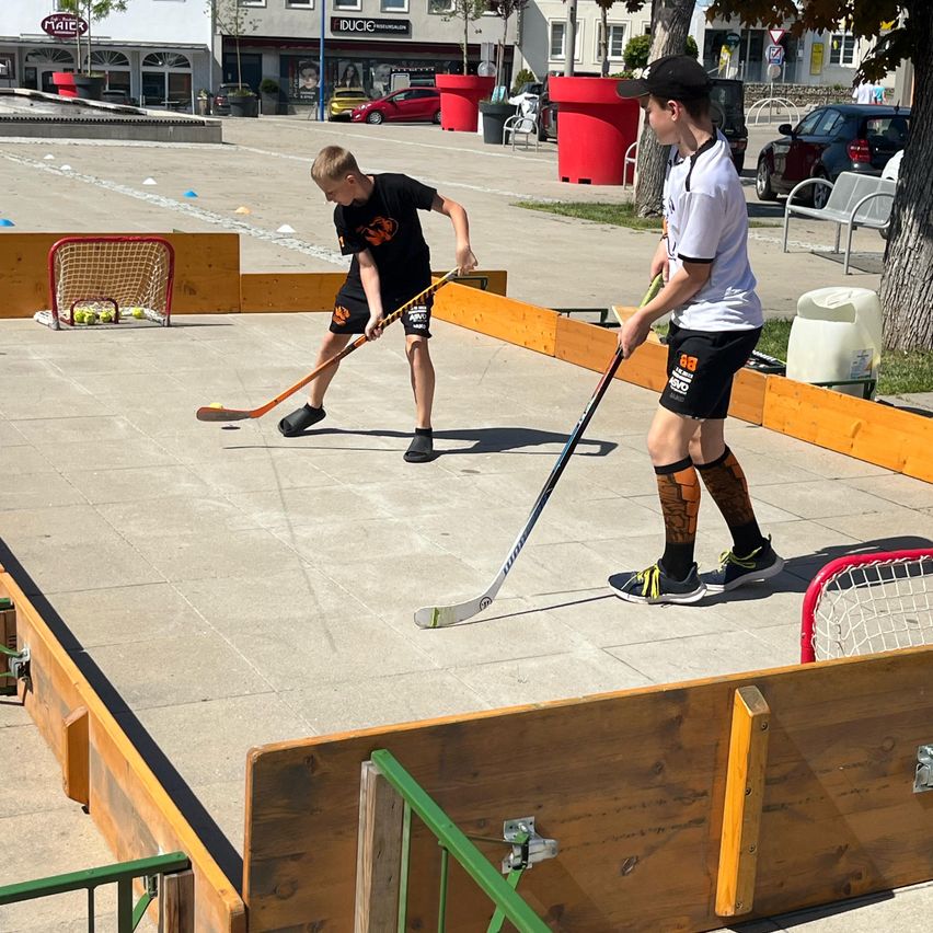 Zwei Jungen spielen Hockey auf einem kleinen, hölzernen Platz in einem offenen Bereich. Ein Junge trägt ein schwarzes T-Shirt und der andere ein weißes T-Shirt. Sie benutzen Hockeyschläger und haben Tore an jedem Ende des Platzes.
