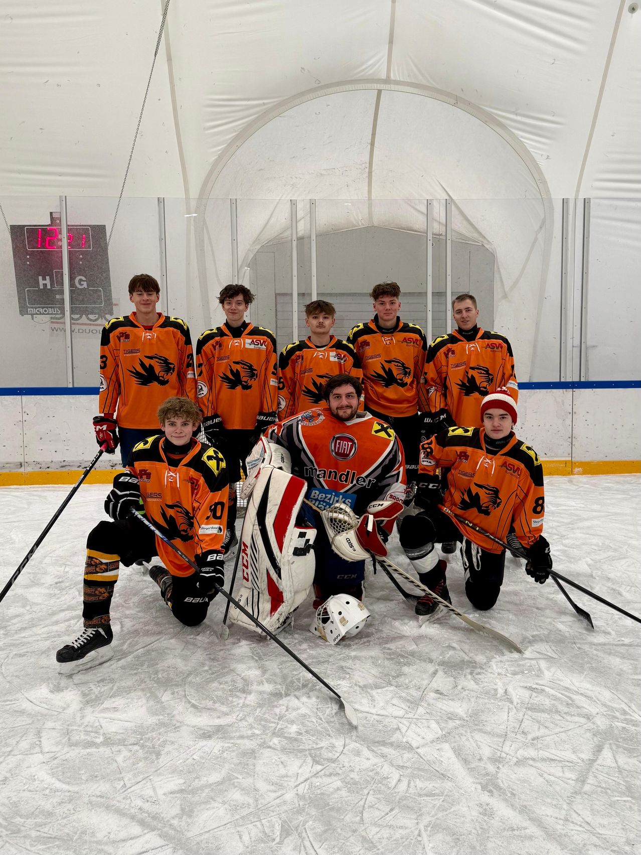 Eine Eishockeymannschaft in orangefarbenen Uniformen posiert für ein Gruppenfoto auf dem Eis, mit einer Anzeigetafel im Hintergrund.