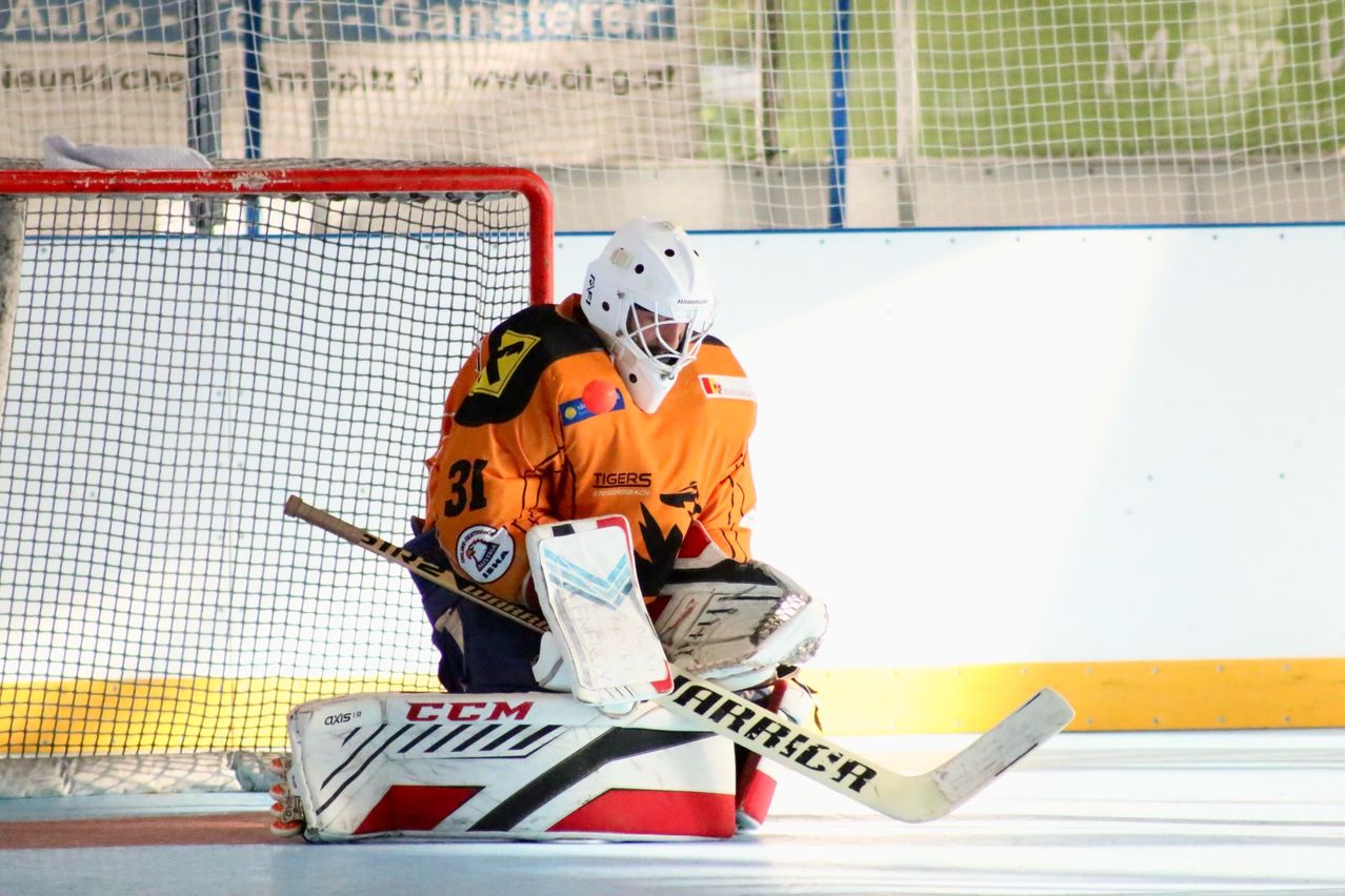 Ein Eishockeytorwart hockt hinter einem Netz und trägt einen Helm und ein orangefarbenes Trikot mit der Aufschrift Tigers. Der Torwart hält einen Eishockeyschläger in der Hand.