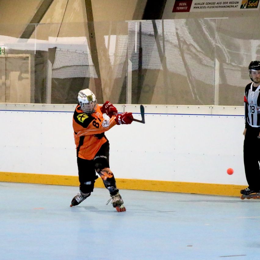 Ein Hockeyspieler in Orange und Schwarz fährt auf Rollschuhen im Innenraum und trifft einen roten Ball mit einem Stock, während ein Schiedsrichter zusieht.