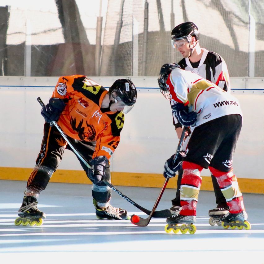 Drei Spieler spielen Rollhockey auf einer Indoor-Eisbahn. Zwei tragen orangefarbene und weiße Trikots, der andere ein schwarzes und weißes Trikot. Sie tragen alle Helme und Handschuhe.