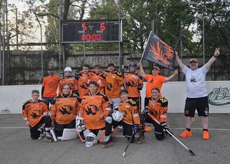 Eine Gruppe von Hockeyspielern in orangefarbenen Trikots und Helmen posiert für ein Foto auf dem Spielfeld. Eine Anzeigetafel hinter ihnen zeigt ein Unentschieden.