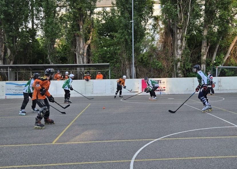 Eine Gruppe Spieler auf Rollschuhen spielt Hockey auf einem Außenfeld. Zuschauer schauen von der Mauer aus zu.