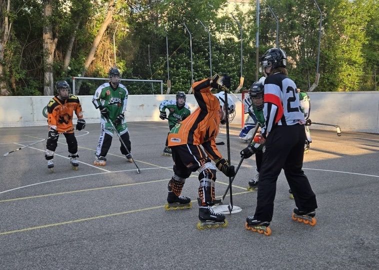 Eine Gruppe von Spielern mit Rollschuhen spielt Hockey auf einer Freiluftbahn. Sie tragen alle Helme, Handschuhe und Rollschuhe. Sie halten Eishockeyschläger und einige von ihnen schauen sich etwas an. Die Bahn hat eine weiße Wand, eine weiße Linie und ein Tor.