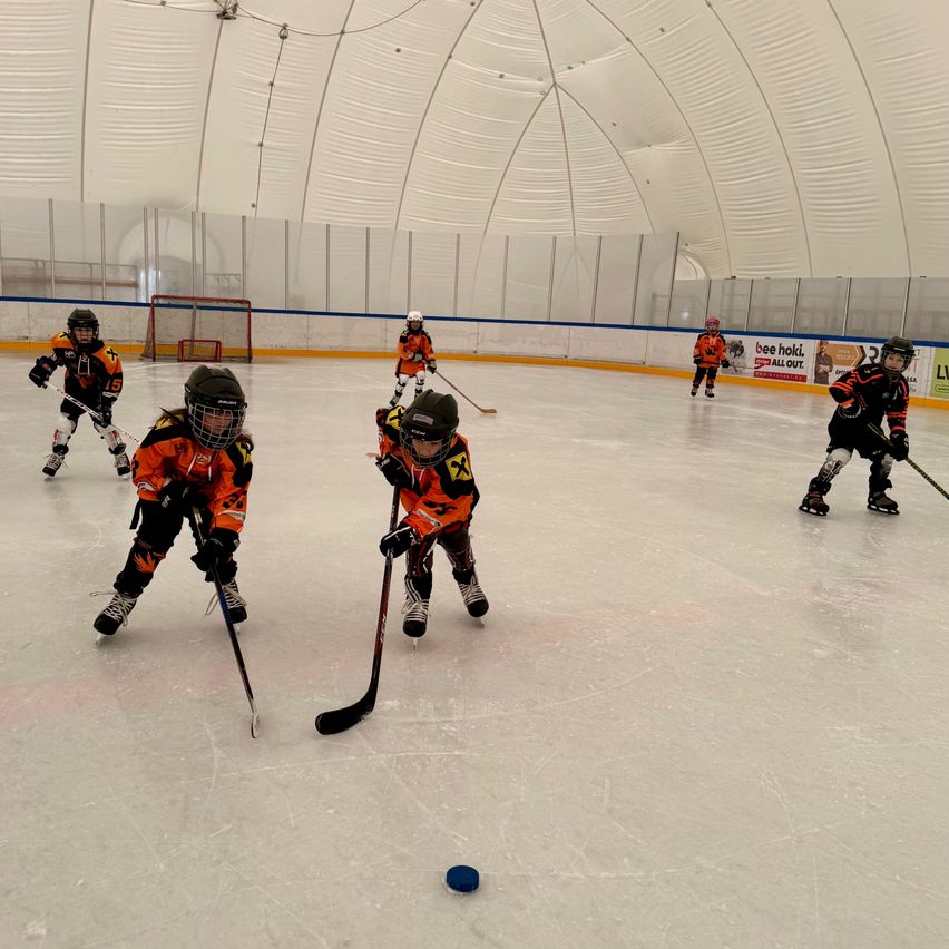 A group of children are playing ice hockey in an indoor ice rink. They are all wearing helmets and hockey gear. The rink has a white ceiling and walls.