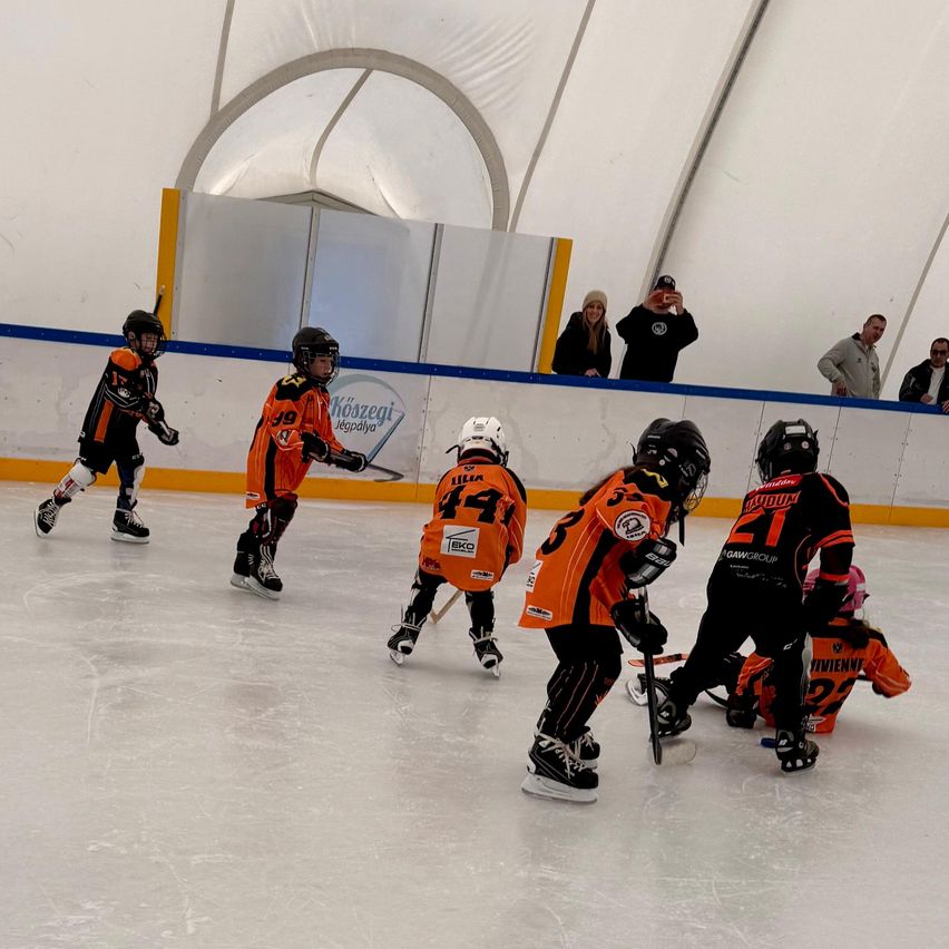 A group of children in orange and black uniforms are playing hockey on an ice rink. A few adults are watching from the sidelines.