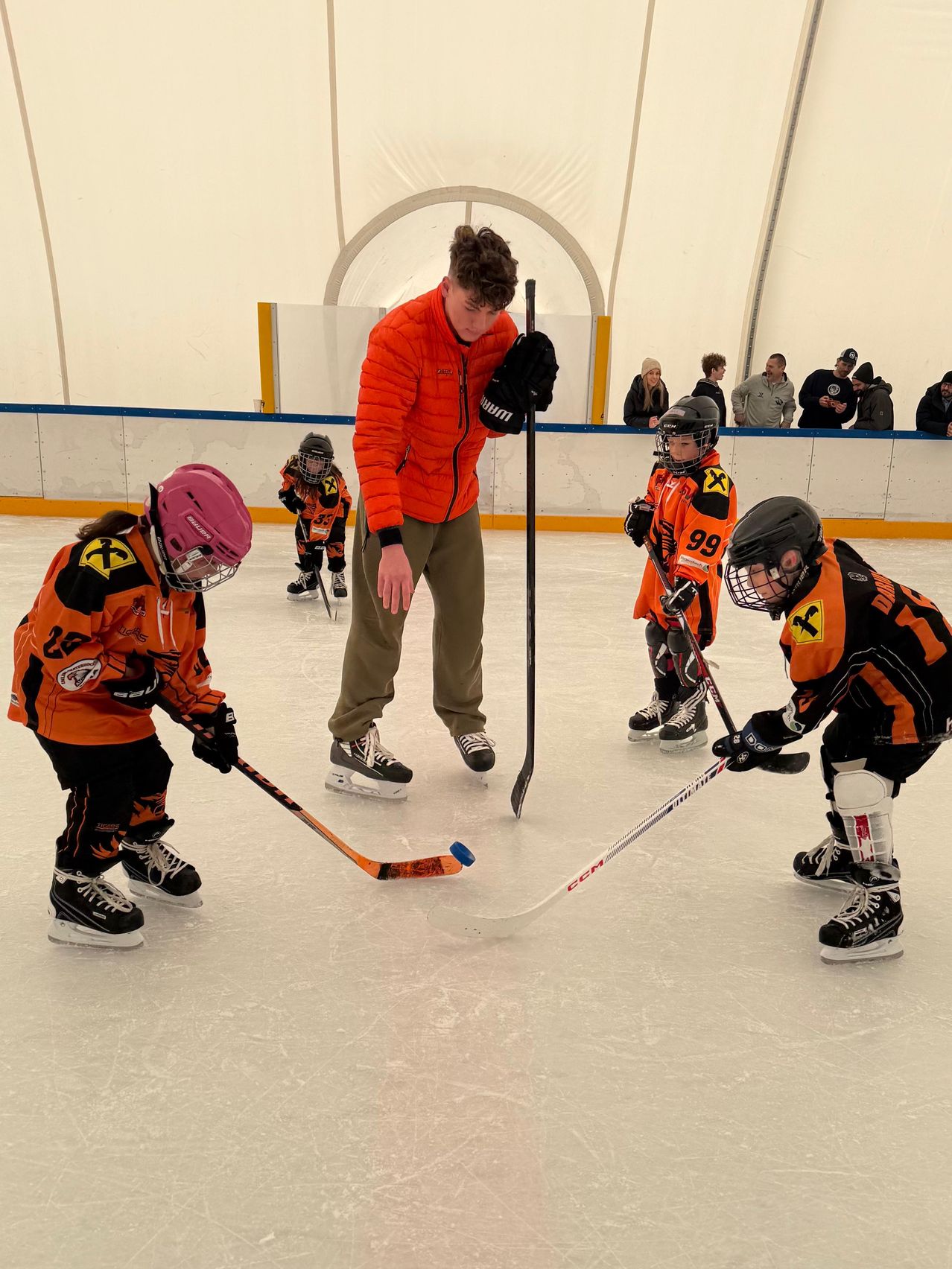 A hockey coach is teaching young players on an ice rink. They wear orange uniforms and helmets. Some are practicing while others watch. The coach holds a hockey stick.