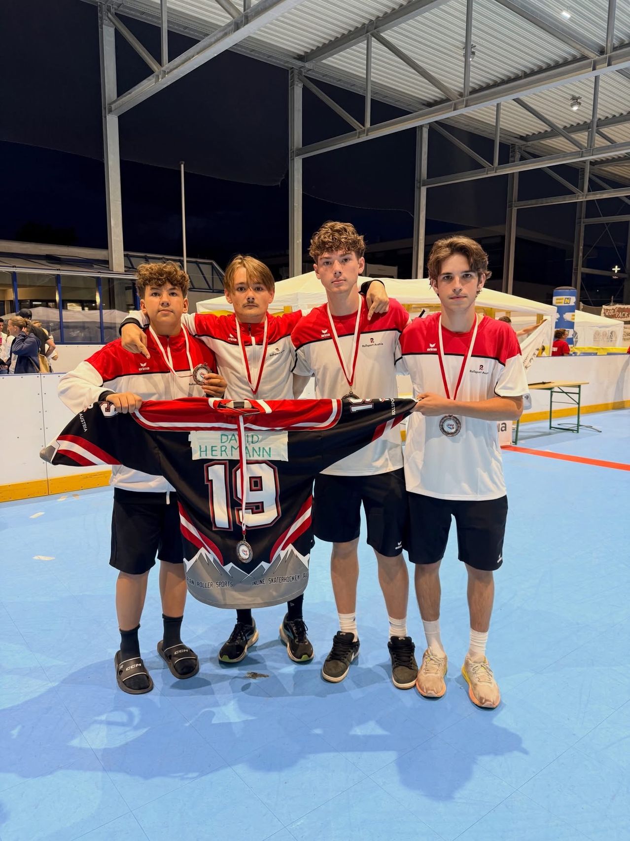 Vier junge Männer in Sporttrikots mit Medaillen um den Hals halten ein Eishockeytrikot mit dem Namen David Hermann und der Nummer 19 hoch. Sie stehen auf einem blauen Boden, wahrscheinlich bei einer Sportveranstaltung.