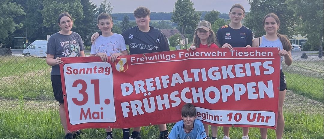Eine Gruppe junger Menschen posiert mit einem Banner vor einem grasbewachsenen Bereich mit Bäumen. Das Banner trägt die Aufschrift 'Dreifaltiger Fruschoppnen' und das Datum '31. Mai'.