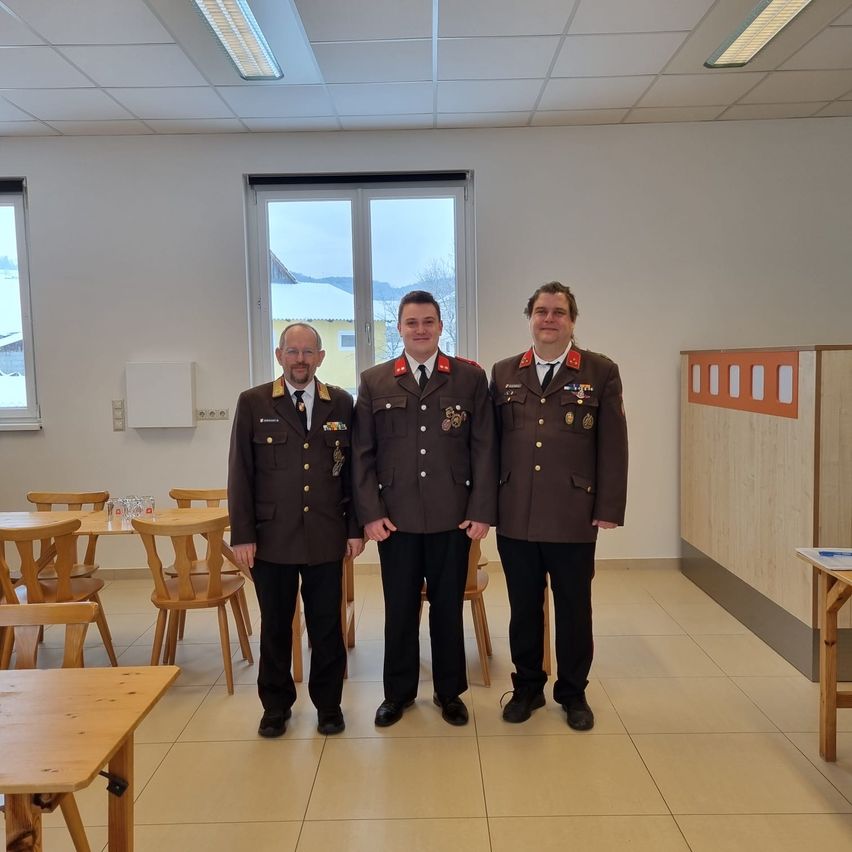 Drei Männer in Militäruniformen stehen in einem Raum mit gefliesten Böden, Holztischen und Stühlen. Der Raum hat große Fenster und eine weiße Wand.