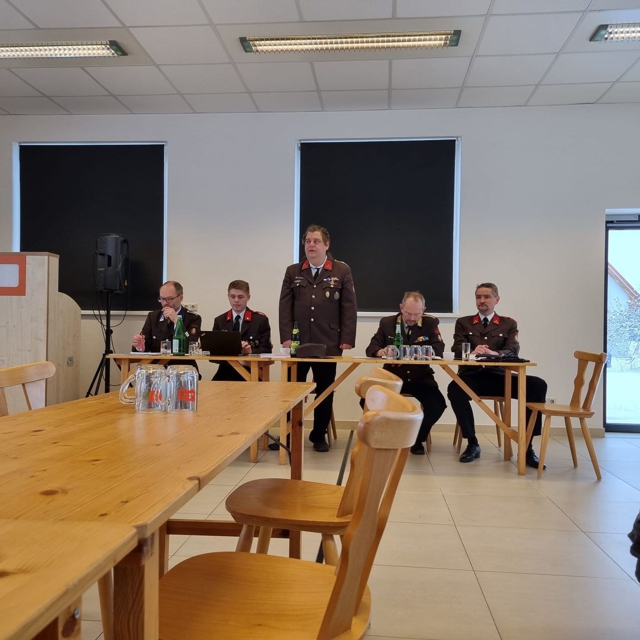 Eine Gruppe von Männern in Militäruniformen sitzt an einem Tisch, einer steht. Ein Laptop und Gläser sind auf dem Tisch. Der Raum hat weiße Wände und gefliesten Boden.