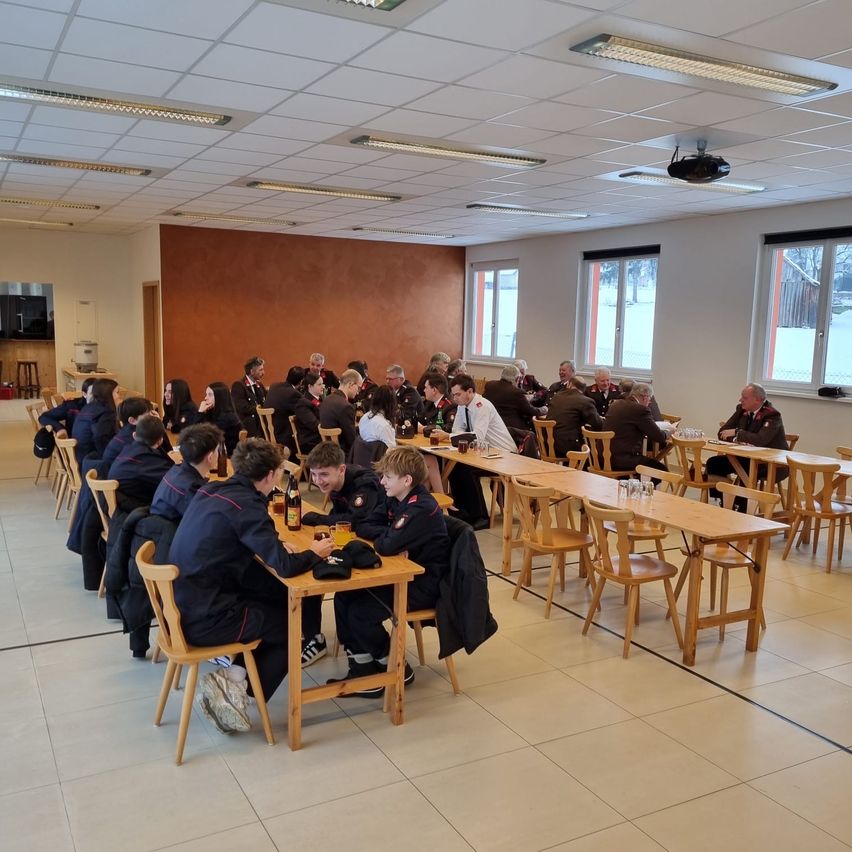 Eine Gruppe von Personen in Uniformen sitzt an Tischen in einem Raum mit weißen Decken, Wänden und einer Holzwand und diskutiert.