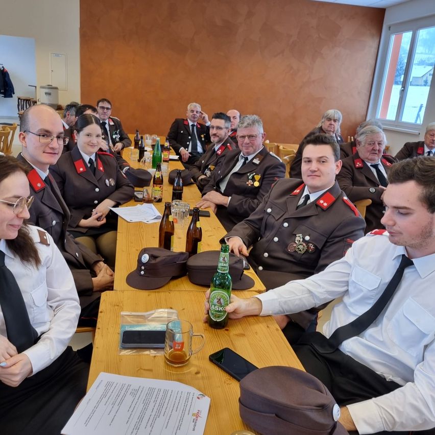 Eine Gruppe uniformierter Personen sitzt um einen langen Tisch. Einige tragen Medaillen und halten Bierflaschen. Ein Mann im Vordergrund hält ein Bier. Papiere und Hüte sind auf dem Tisch.