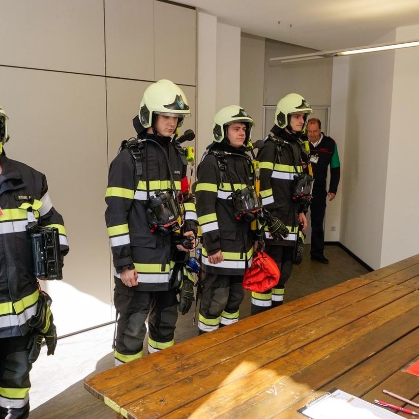 Eine Gruppe von Feuerwehrleuten steht in einem Raum mit Helmen, hält Ausrüstung und trägt Rucksäcke. Eine Person hält eine Kamera. Der Raum verfügt über einen Holztisch, und ein Mann geht hinter ihnen.