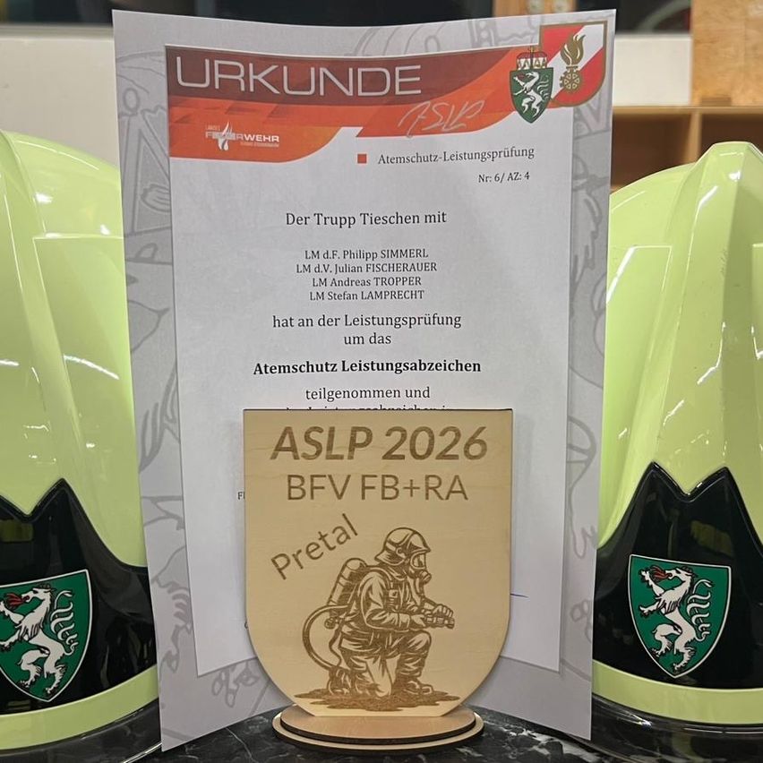 Ein Zertifikat auf einem Ständer zeigt die Namen von Teammitgliedern, die einen Atemschutzleistungstest absolviert haben. Das Zertifikat zeigt ein Logo und den Text 'Atmschutz-Leistungsprüfung', sowie eine Plakette mit dem Text 'AStP 2026 BFV FB+RA Pretal'. Hinter dem Zertifikat stehen zwei grüne Helme mit einem Löwenwappen.