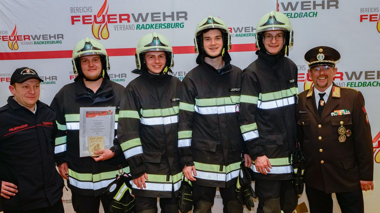 Vier Feuerwehrleute stehen vor einem Banner mit den Worten 'Feuerwehr Verband Radkersburg'. Sie tragen schwarze und grüne Uniformen mit reflektierenden Streifen. Der Mann auf der linken Seite hält ein Zertifikat. Der Mann in der Mitte lächelt, während die anderen nach vorne schauen.
