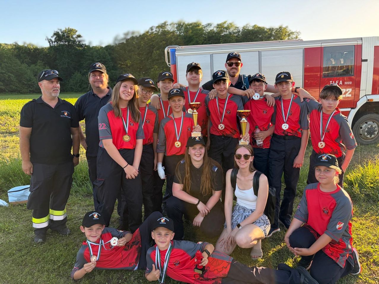 Eine Gruppe junger Feuerwehrleute, sowohl Männer als auch Frauen, in roten und grauen Uniformen, posiert mit ihren Trophäen auf einer Grasfläche vor einem Feuerwehrauto.