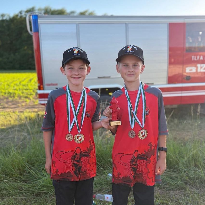 Zwei junge Jungen stehen in einem Feld mit Medaillen um den Hals, lächelnd und halten eine Trophäe. Ein roter LKW mit Text ist im Hintergrund.