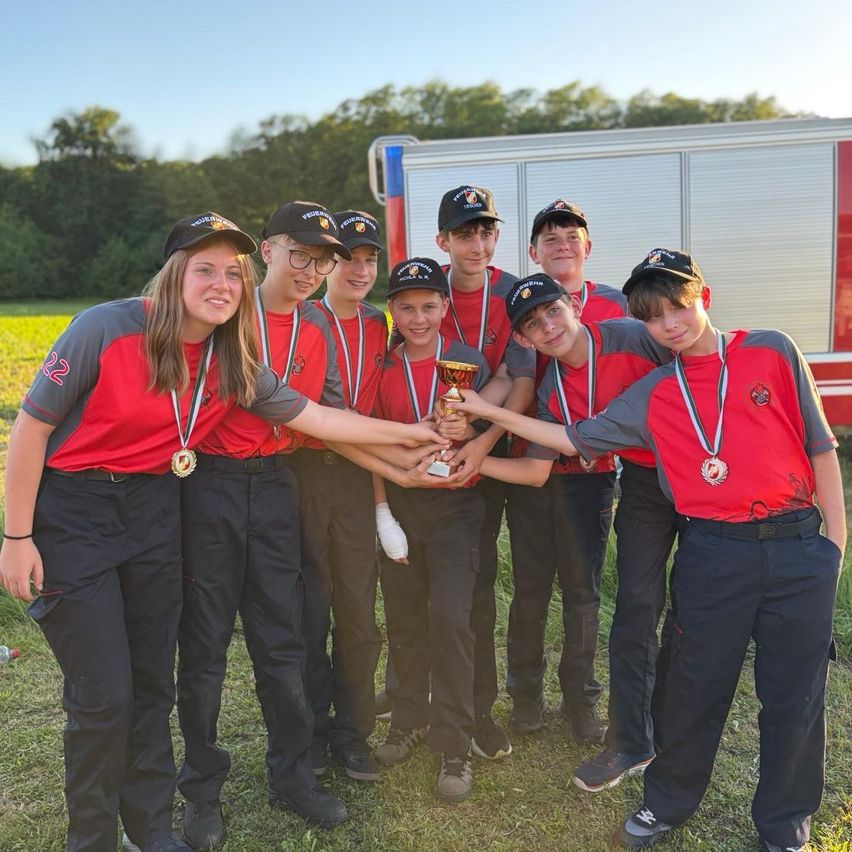 Eine Gruppe junger Athleten in gleichen Uniformen mit Medaillen um den Hals und einem Pokal in den Händen, posiert für ein Foto auf einem Feld mit einem Feuerwehrauto im Hintergrund.