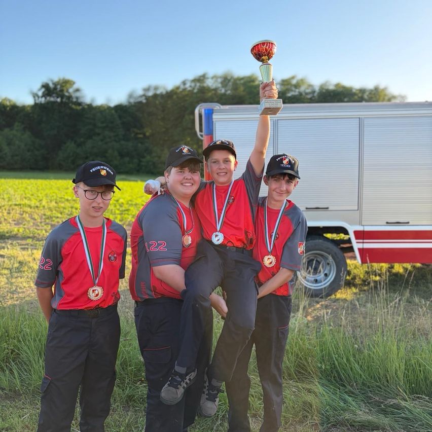 Vier junge Personen posieren mit Medaillen und einem Pokal für ein Foto. Sie stehen auf einem Feld mit einem Feuerwehrfahrzeug im Hintergrund.