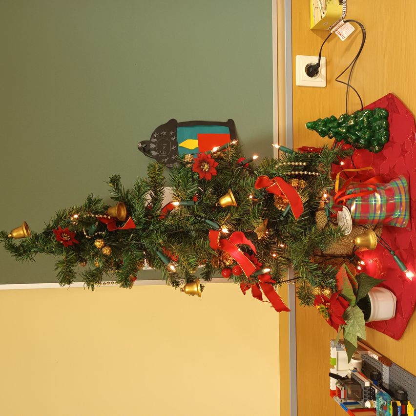 Ein dekorierter Weihnachtsbaum mit roten Bändern, Ornamenten und einem grünen Baumschmuck steht auf einem roten Teppich an einer grünen Wand.