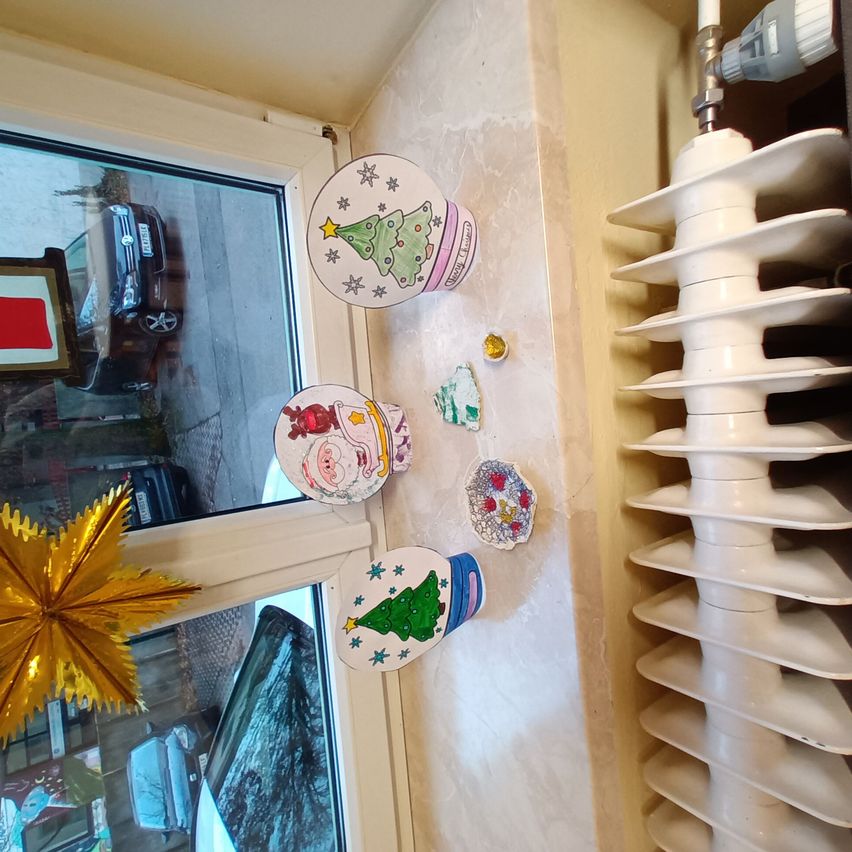 Ein Fenster mit Weihnachtsdekoration, einschließlich eines Weihnachtsbaums, des Weihnachtsmanns und Ornamenten, befindet sich neben einem weißen Heizkörper. Draußen ist ein Auto auf der Straße geparkt.