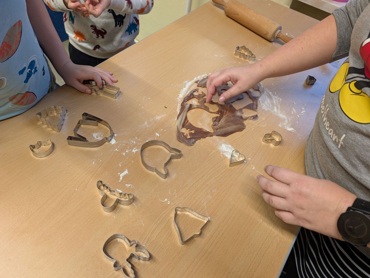Mehrere Kinder stellen Kekse mit Ausstechern auf einem Holztisch her. Der Tisch ist mit Mehl und Ausstechern übersät. Ein Kind schneidet Teig aus.