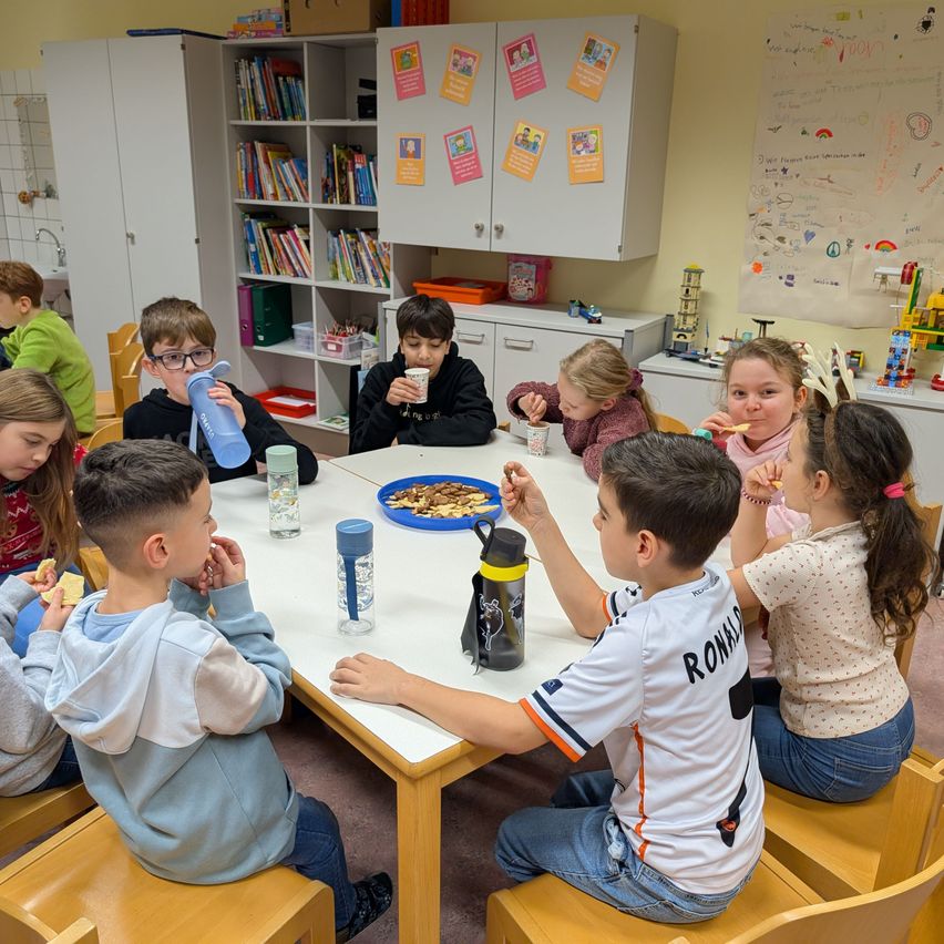 Eine Gruppe von Kindern sitzt in einem Klassenzimmer an einem Tisch und isst Snacks und trinkt aus Bechern. Der Tisch hat einen Teller mit Snacks und Wasserflaschen. Das Klassenzimmer hat Bücherregale, Schränke und eine Tafel.