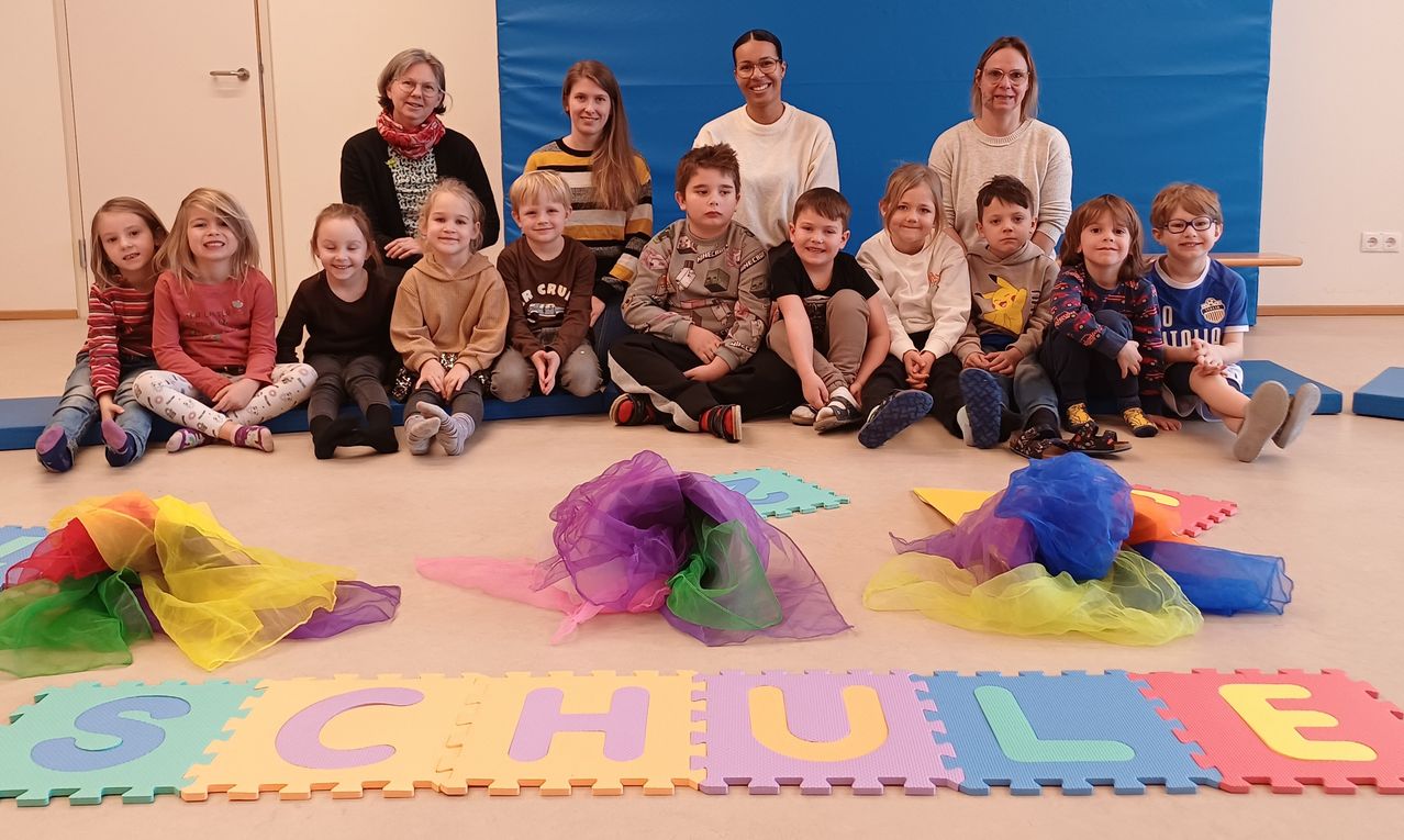 Eine Gruppe von Kindern und Erwachsenen in einem Raum. Sie sitzen auf dem Boden vor einer blauen Wand. Die Kinder lächeln und tragen verschiedenfarbige Kleidung. Sie sind auch von farbenfrohen Matten mit dem Wort 'SCHULE' umgeben.