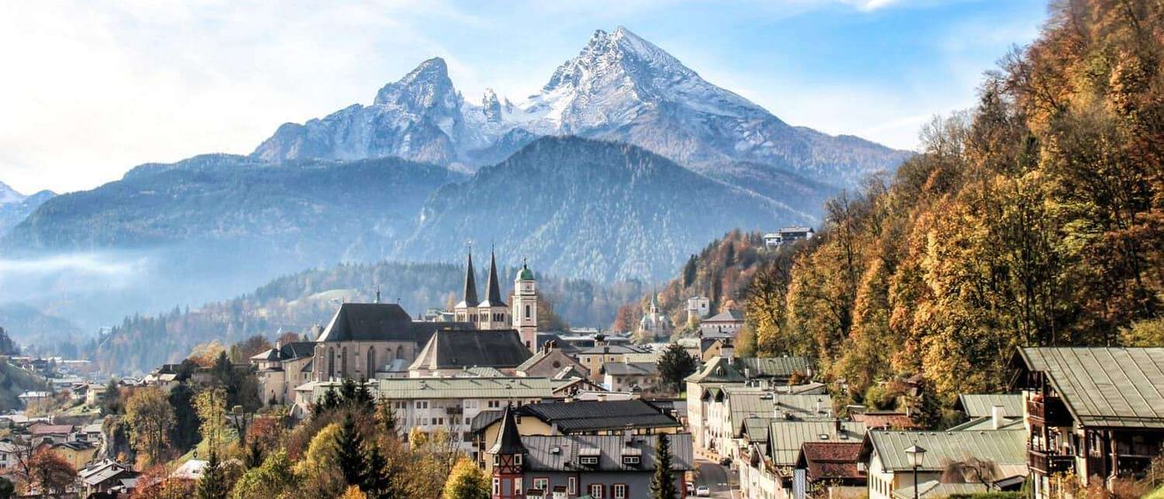 Ein malerisches Städtchen, eingebettet unter schneebedeckten Bergen unter einem teilweise bewölkten Himmel, mit herbstlichen Bäumen und historischen Gebäuden.