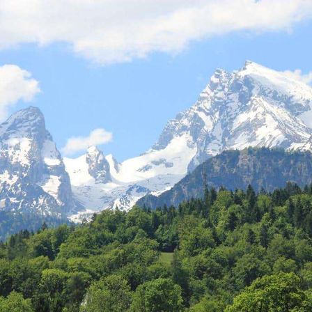 Eine majestätische Bergkette mit schneebedeckten Gipfeln und üppigem Grün unter einem teilweise bewölkten blauen Himmel.