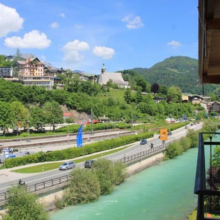 Ein Blick von einem Balkon zeigt eine Stadt mit Bergen, einem Fluss und einer Straße mit Autos und Motorrädern. Bäume und Büsche säumen den Fluss, und ein Bahngleis verläuft daneben.
