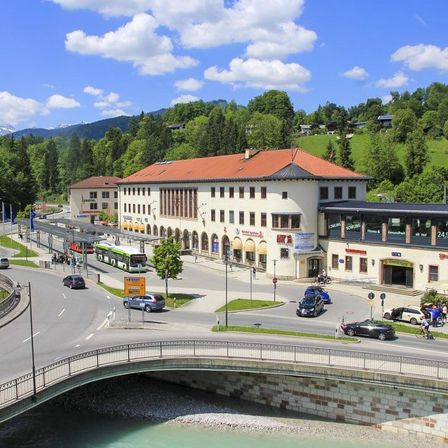 Ein Luftbild einer belebten Stadt mit einem Fluss, Autos und einer Brücke. Gebäude säumen die Straße mit einer Mischung aus moderner und traditioneller Architektur. Bäume und Berge sind im Hintergrund zu sehen.