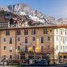 A yellow hotel named Schwabenwirt with several windows is situated in front of a snowy mountain. There are a few shops below the hotel. A person is walking on the street.