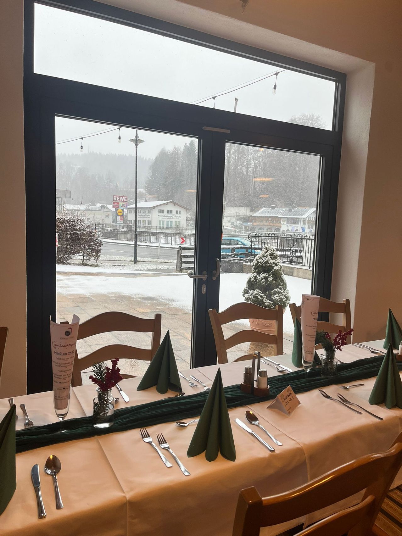 A dining table set with silverware, napkins, and flowers is inside a room with large windows. The view outside shows a snowy road and a car.