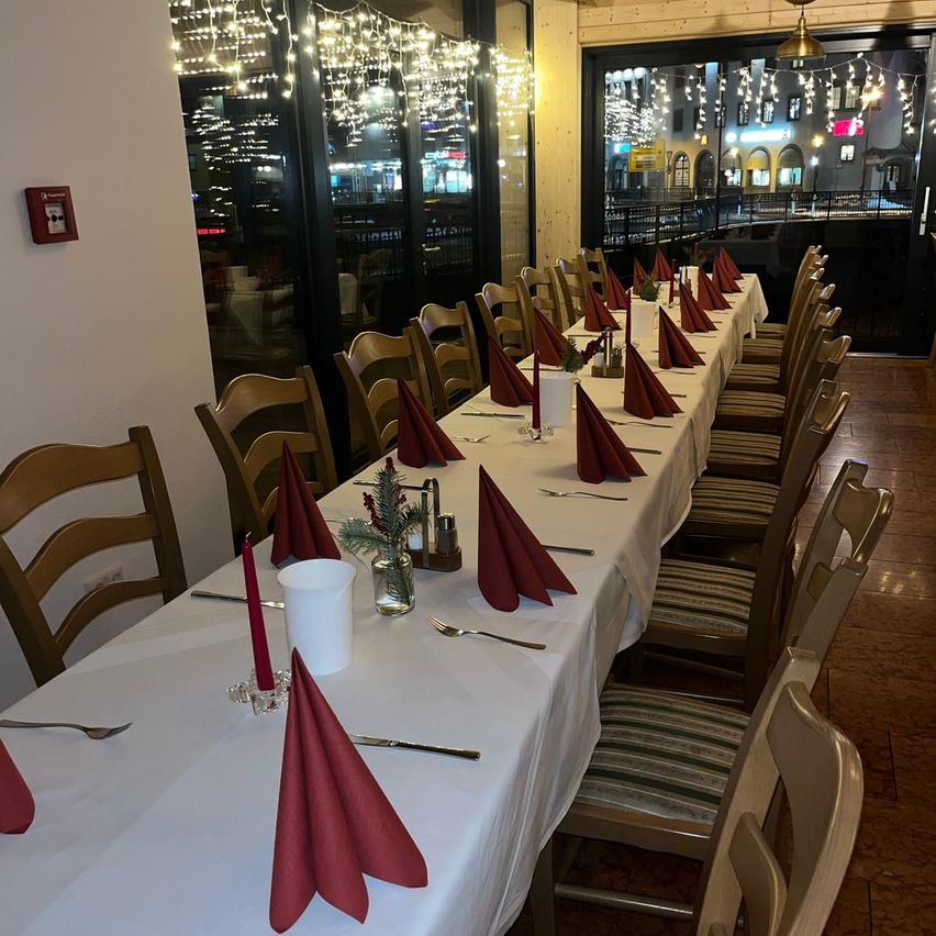 A restaurant dining area set up for a large gathering with a long table covered in white cloth, adorned with red napkins, candles, and cutlery. The room has large windows showing a city view at night.